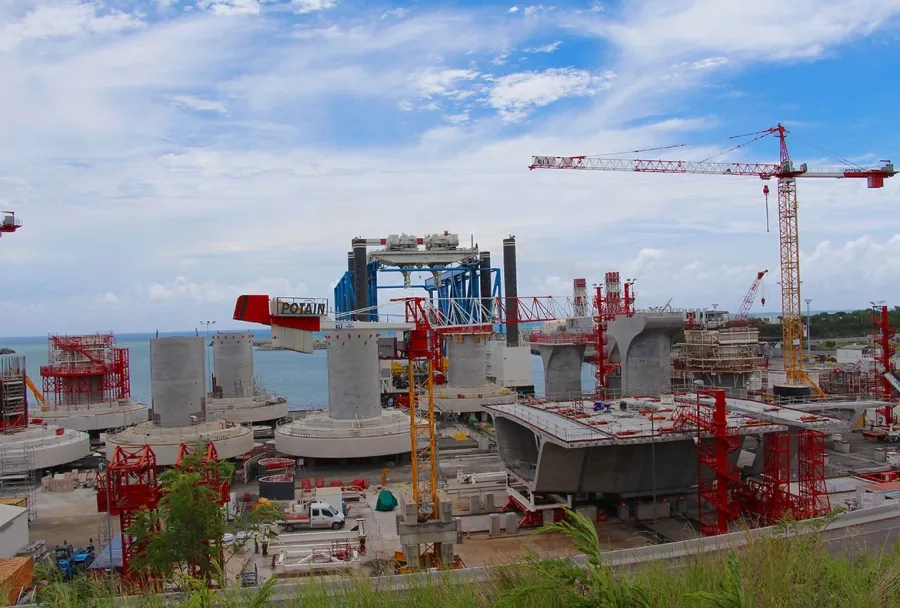 Une fois leur phase de maturation terminée, les embases et têtes de piles sont embarquées directement depuis l’aire de préfabrication sur la barge (au fond en bleu) qui les mettra en place. Une fois leur phase de maturation terminée, les embases et têtes de piles sont embarquées directement depuis l’aire de préfabrication sur la barge (au fond en bleu) qui les mettra en place.