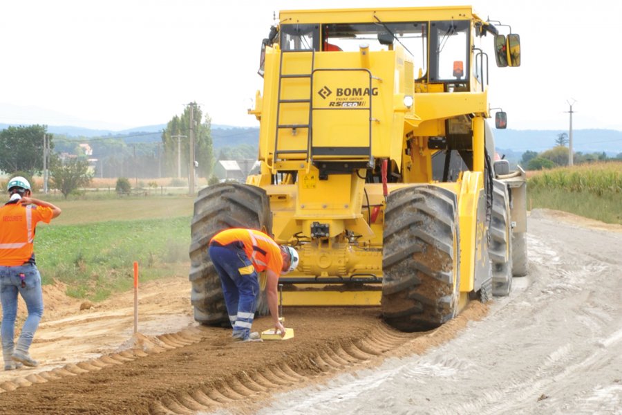 Derrière le malaxeur Bomag RS 650, un technicien vérifie visuellement l'homogénéité du mélange entre le matériau de l’ancienne chaussée et le liant hydraulique, et un autre mesure la teneur en eau du matériau traité.