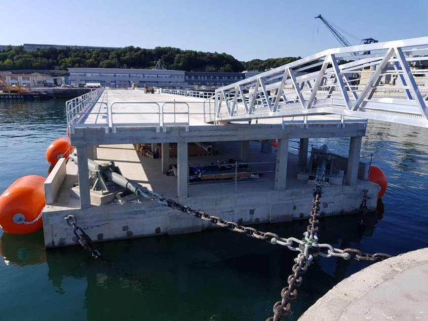 Les chaînes d’amarrage du ponton (ici les deux chaînes côté musoir) sont fixées à celui-ci via des amortisseurs (en gris) dimensionnés pour reprendre les efforts d’accostage et d’amarrage des frégates. Pour supporter ces charges élevées, les amortisseurs reposent sur des socles en béton très fortement armé. 