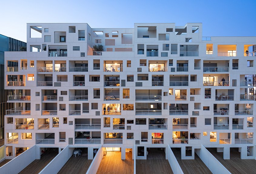 L’opération combine des logements, enserrés dans une résille porteuse en béton blanc autoplaçant, des bureaux, enveloppés par un mur rideau en aluminium, et des commerces en rez-de-chaussée.