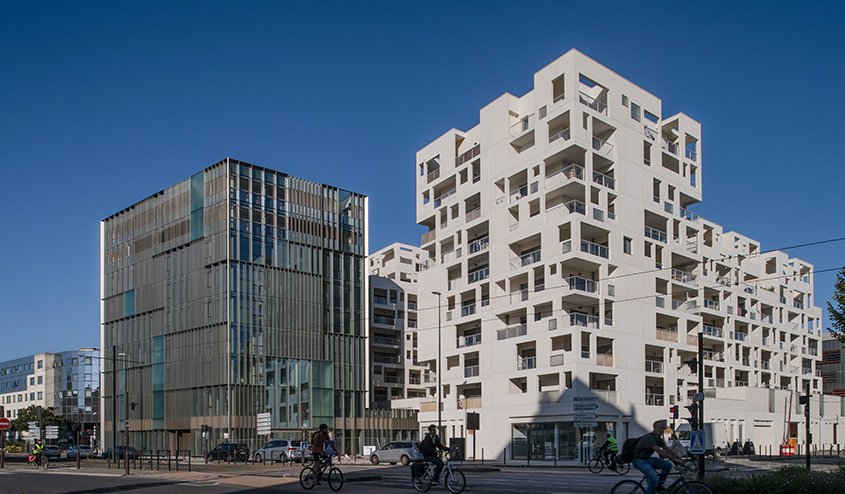 L’opération combine des logements, enserrés dans une résille porteuse en béton blanc autoplaçant, des bureaux, enveloppés par un mur rideau en aluminium, et des commerces en rez-de-chaussée.